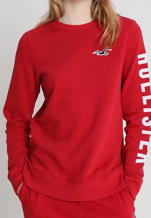 Kvinde iført en rød langærmet sweatshirt med "HOLLISTER" i hvide bogstaver på ærmet og et lille fuglelogo på brystet.