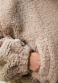 Hyggelig beige fleece-pullover med en blød, tekstureret overflade og stort manchet-detalje, der har en skjult lomme foran.