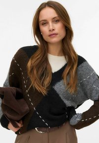 Navaden pulover z veliko pletenino z vzorcem črnih, sivih in rjavih diamantov; vsebuje bele šive kot poudarke. Model drži rjavo torbo.