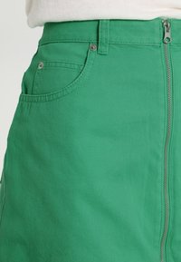 Falda de mezclilla verde con cierre frontal de cremallera, dos bolsillos laterales y tela texturizada con un acabado suave.