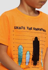 Oranje katoenen T-shirt met een grafisch ontwerp van verschillende skateboardontwerpen in groen, blauw en zwart, gemarkeerd met afmetingen en "SKATE THE UNKNOWN."