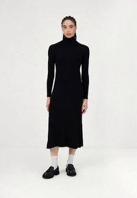 Juoda kosztuota midi ilgio suknelė aukštu kaklu, ilgomis rankovėmis, aptempianti siluetą ir šiek tiek platėjanti ties apačia. Dėvima su juodais batais ir baltais kojinėmis.