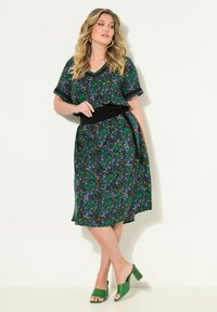 Robe fleurie noire avec des motifs verts et violets, manches courtes, décolleté en V, et une ceinture noire à la taille. Associée à des mules à talons épais verts.