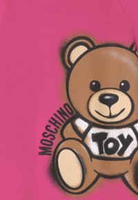Nallebjörnsgrafik med "Toy" på sin tröja och "MOSCHINO"-text på ljusrosa tygbakgrund.