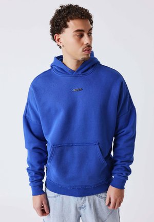 Jonge man met krullend haar, gekleed in een felblauwe hoodie en lichte spijkerbroek, kijkt naar links tegen een effen achtergrond.