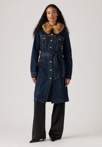 Manteau en denim avec un col en fausse fourrure, couleur bleue foncée, ceinture à la taille, fermeture à boutons et poches. Manches longues et design mi-long.