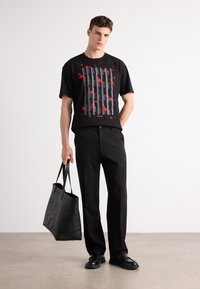 Schwarzes T-Shirt mit einem roten und grauen Vogelmotiv, kombiniert mit schwarzen Hosen und einer schwarzen Leder-Tote-Tasche mit geprägtem Logo.