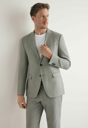 Mężczyzna w jasnoszarym garniturze i białej koszuli poprawiający kołnierz marynarki, stojący na tle gładkiej białej ściany, patrzący na bok.