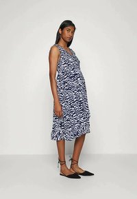 Mulher a usar um vestido midi sem mangas, com padrão em azul marinho e branco, com bainha em babados e sapatos planos pretos com atilhos no tornozelo, de pé contra um fundo simples.