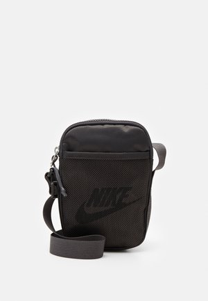 Mała czarna torebka Nike na ramię z siatkową przednią kieszenią, zapięciem na zamek i regulowanym paskiem na białym jednolitym tle.