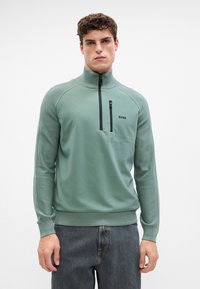 Grüner Reißverschluss-Pullover aus glattem Material mit gerippten Bündchen und Saum. Verfügt über eine Brusttasche und ein kleines schwarzes Logo an der Seite.