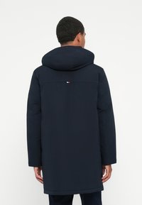 Mann in einer marineblauen Kapuzenparka, der von der Kamera wegsteht und vor einem einfarbigen weißen Hintergrund steht.