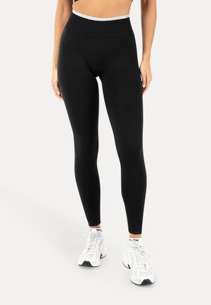 Kvinde iført sort leggings med høj talje og hvide sneakers, stående mod en ensfarvet lys baggrund.