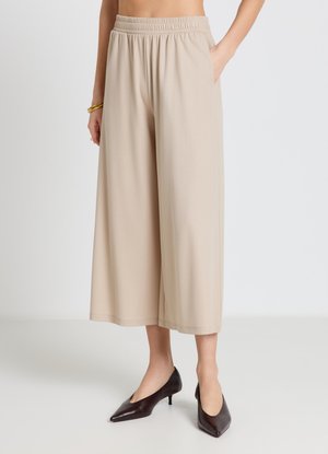 Beige wide-leg cropped pants with elastic waistband, worn with dark brown pointed-toe low heels and gold bracelets on the left wrist.