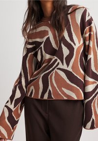 Stickad tröja med ett djärvt abstrakt mönster i brun, kräm och rost, med båtformad halsringning och vida ärmar, i kombination med bruna byxor.