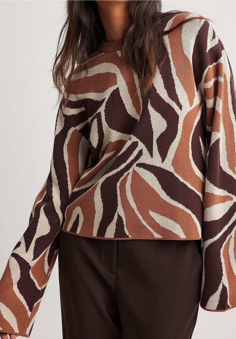 Stickad tröja med ett djärvt abstrakt mönster i brun, kräm och rost, med båtformad halsringning och vida ärmar, i kombination med bruna byxor.