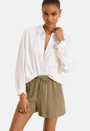 Vrouw die een wit, lang mouw knoophemd draagt en olijfgroene shorts met trekkoord, poserend met één hand bij haar nek en de andere langs haar zijde.