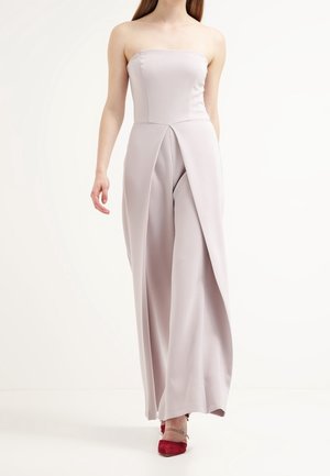 Vrouw draagt een strapless jumpsuit in lichtlavendel met brede pijpen en een voorpaneel, gecombineerd met rode puntige hoge hakken, tegen een effen witte achtergrond.