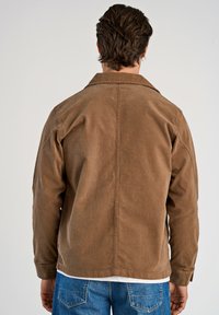 Brun kordflorsjacka med krage, synliga sömnadsdetaljer och en något avslappnad passform, matchad med blå denimjeans.