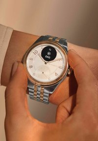 Silberne und goldfarbene Smartwatch mit einem runden weißen Ziffernblatt, großen Zahlen und einer auf dem Bildschirm angezeigten EKG-Funktion. Metallarmband mit Gliedern.