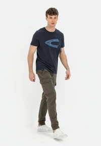 Marineblauwe katoenen t-shirt met een gestructureerd blauw logo, gecombineerd met olijfgroene cargobroek met zakken, en witte sneakers met grijze accenten.
