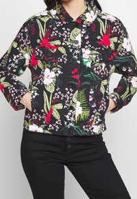 Giacca di jeans con design floreale su base nera, caratterizzata da fiori rossi e bianchi, foglie verdi, tasche frontali e chiusura a bottoni a pressione.
