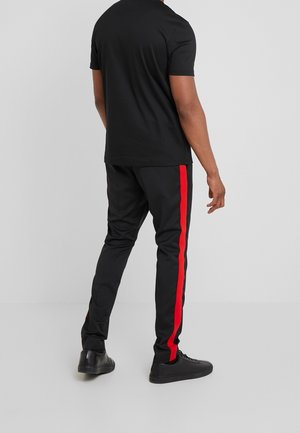Camiseta negra de manga corta combinada con pantalones negros que tienen una raya roja vertical a lo largo del costado. Lleva zapatillas negras.