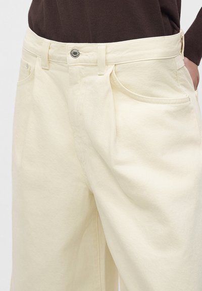 Pantalon taille haute couleur crème avec poches avant et fermeture par bouton, porté avec un haut marron foncé, visible de la taille jusqu'à mi-cuisse.