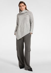 Maglione grigio chiaro lavorato a maglia con collo a scialle drappeggiato e orlo diagonale, caratterizzato da un motivo a trecce testurizzate. Abbinato a pantaloni grigi scuri a gamba larga.