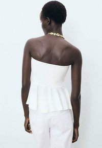 White strapless peplum top with a fitted bodice, made from smooth fabric. Worn with light-colored pants and complemented by a gold necklace.
