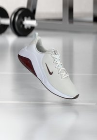 Valkoiset verkkomaiset urheilukengät punaisilla yksityiskohdilla, pyöreä kärki ja koristeellinen nauhajärjestelmä. Nike-logo ruskeana sivussa ja kielen päällä.