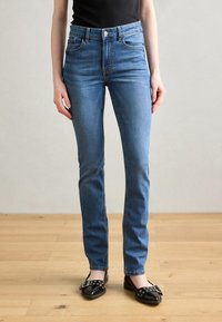 Jeans din denim albastru clasic cu talie înaltă, cinci buzunare și o croială ajustată, asortați cu pantofi plat negri cu accente metalice.
