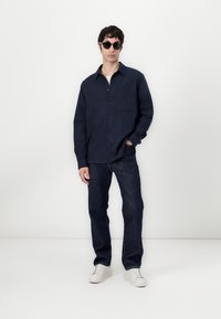 Chemise bleu marine à boutons avec une poche poitrine, associée à un jean bleu foncé et des baskets blanches. Le modèle porte des lunettes de soleil noires.