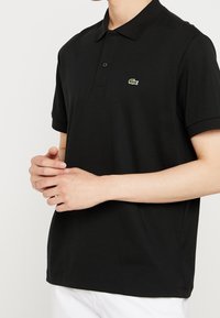 Polo-shirt noir en tissu coton, avec col, manches courtes et un petit logo Lacoste vert brodé sur la poitrine.