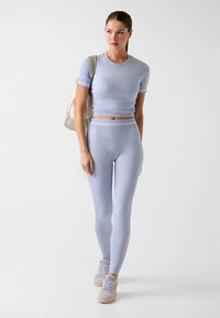 Hellblaues sportliches Crop-Top und Leggings aus elastischem Stoff. Das Outfit hat ein eng anliegendes Design, kombiniert mit beigen Turnschuhen und einer hellen Tasche.