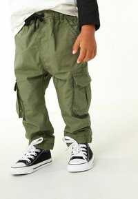 Pantaloni cargo verdi con polsini elasticizzati, dotati di tasche anteriori e laterali, abbinati a sneaker nere con suole e lacci bianchi.