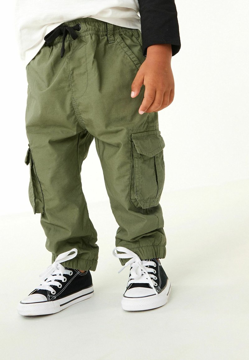 Pantaloni cargo verdi con polsini elasticizzati, dotati di tasche anteriori e laterali, abbinati a sneaker nere con suole e lacci bianchi.
