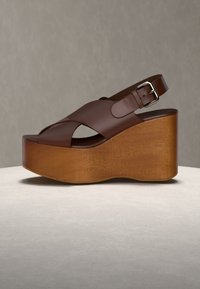 Sandale compensée en cuir marron avec talon compensé en bois et bride à boucle argentée sur fond neutre.