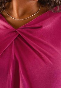 Fuchsiový top s designem zkrouceného uzlu na výstřihu, vyrobený z hladké, měkké látky. Obsahuje jemný stříbrný náhrdelník.