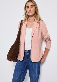 Světle růžový blazer z jemné látky, s elegantním střihem, dvěma předními kapsami a jedním knoflíkem, v kombinaci s modrými džínovými kalhotami.