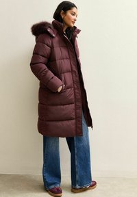 Bordowa kurtka puchowa z futrzanym kapturem, pikowany design, guziki z przodu i boczne kieszenie, zestawiona z dżinsami o szerokich nogawkach i tenisówkami.
