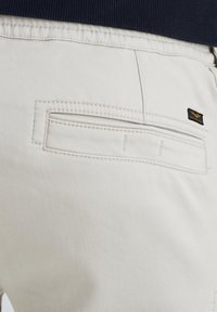 Light beige pants with a back pocket featuring topstitching. A small black label is attached to the side, adding a detail to the design.