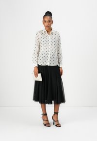 White, polka-dotted blouse with a button-down design, paired with a black, tulle skirt. Accessories include a textured white clutch and black heels.