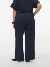 Persoon draagt marineblauwe wijde broek en bijpassende top, staat met één hand op de heup en zwarte schoenen op een witte achtergrond.