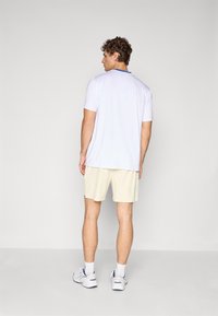Cămașă sport albă cu mânecă scurtă și guler albastru, combinată cu pantaloni scurți bej deschis și adidași albaștri. Material neted. Vedere din spate.