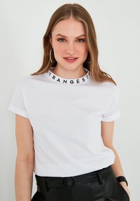Hvid bomuld t-shirt med oprullede ærmer og en sort trykt krave, der står "STRANGERS." Båret med højttlivede sorte bukser.