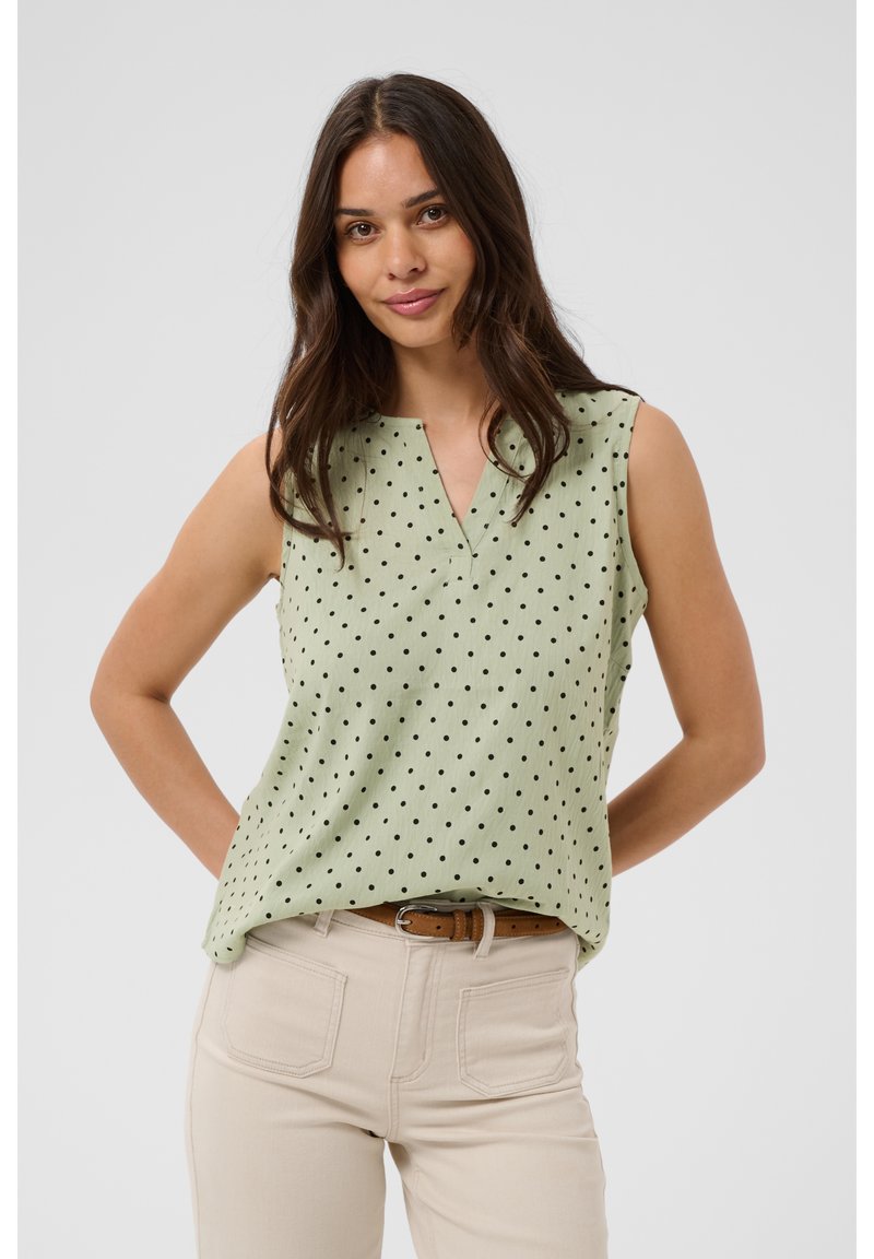 Femme aux longs cheveux foncés portant un haut sans manches vert clair à pois noirs et un pantalon beige avec une ceinture marron.