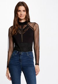 Black long-sleeve top featuring lace and polka dot mesh accents, a high neck, and a fitted body, paired with blue high-waisted jeans.