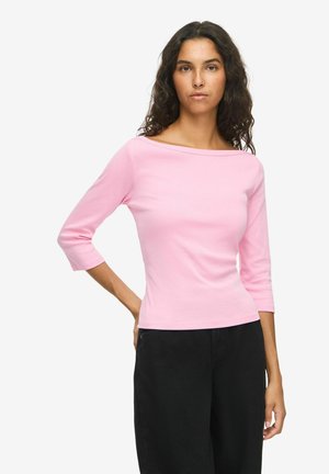 Femme aux cheveux bouclés et foncés portant un top rose clair à manches trois-quarts et un pantalon noir, debout avec une main sur la hanche.