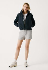 Marineblaue, mit Reißverschluss versehene Jacke mit Kapuze, weiße Logodetails, kombiniert mit grauen Shorts und hellen Sneakers. Glatte Textur und cropped Design.